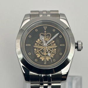 May include: A silver wristwatch with a dark grey face. The watch face displays a gold emblem with a crown, wings, and other symbols. It has a silver strap and a side dial.