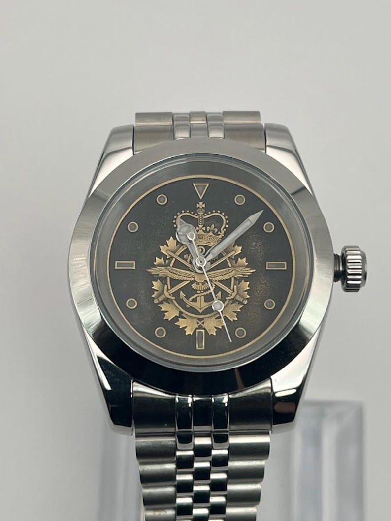 May include: A silver wristwatch with a dark grey face. The watch face displays a gold emblem with a crown, wings, and other symbols. It has a silver strap and a side dial.