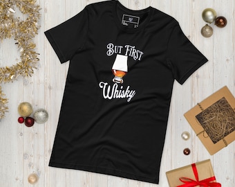 May include: Black t-shirt with the text "But First Whisky" in white, along with a graphic of a whisky glass. The shirt is displayed on a light wood surface, with holiday decorations in the background.
