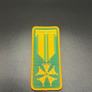 May include: A rectangular patch with a green background and a yellow border. The patch features a vertical design with two yellow stripes and a cross-shaped emblem. The text "VIERDAAGSE NIJMEGEN" is at the top.