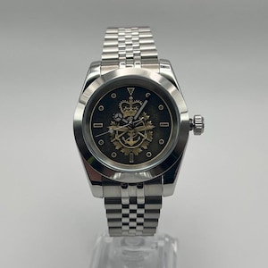 May include: A silver wristwatch with a stainless steel strap. The watch face is dark with a gold emblem featuring a crown, anchor, and other symbols. The watch has a round face and a textured bezel.