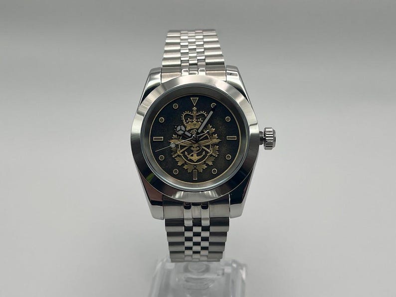 May include: A silver wristwatch with a stainless steel strap. The watch face is dark with a gold emblem featuring a crown, anchor, and other symbols. The watch has a round face and a textured bezel.