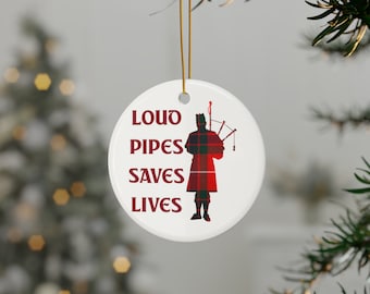 May include: A round, white ceramic Christmas ornament featuring the text "LOUD PIPES SAVES LIVES" in maroon. A silhouette of a bagpiper in a red and green plaid kilt is on the right. It hangs from a gold string, ideal for festive decorations.