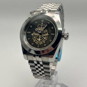 May include: A silver wristwatch with a black face, showcasing a gold crown and anchor design. The watch has a stainless steel strap and a round face with small circular markers. It is displayed on a clear stand.