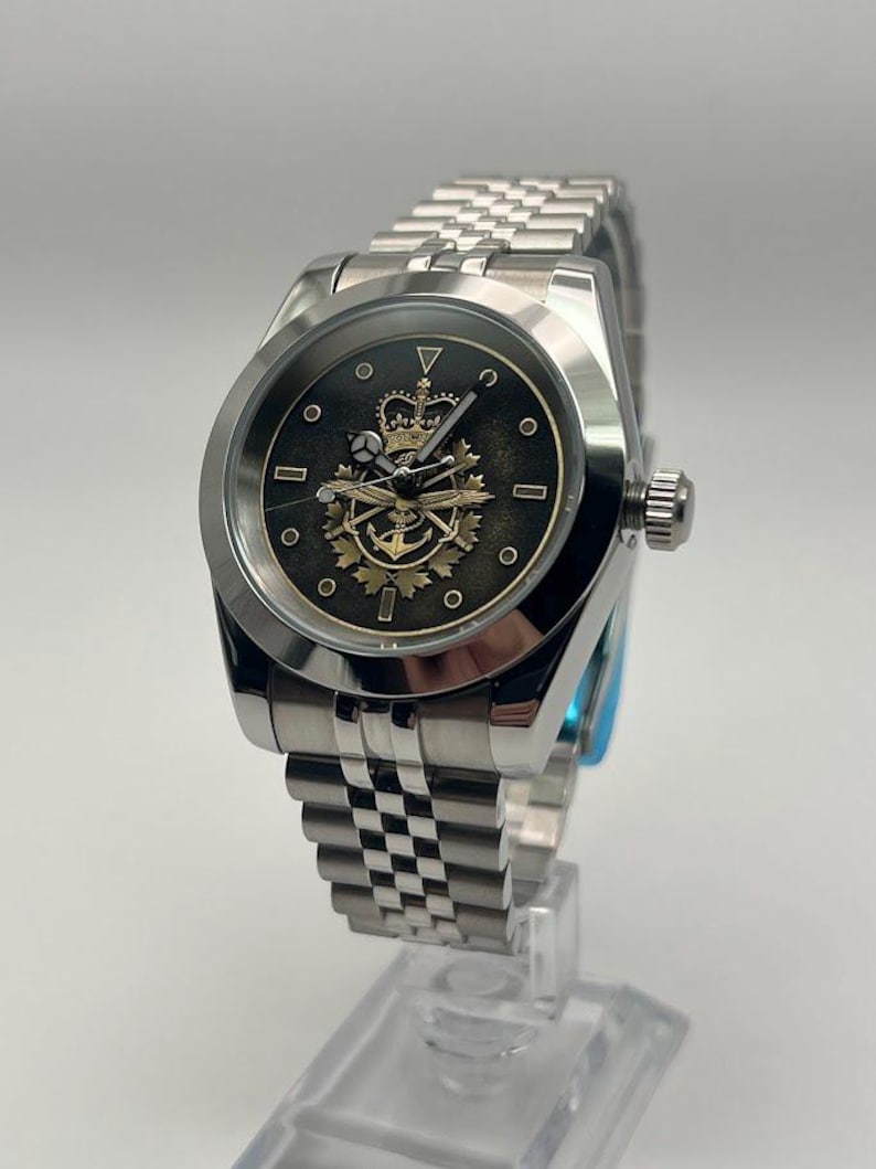 May include: A silver wristwatch with a black face, showcasing a gold crown and anchor design. The watch has a stainless steel strap and a round face with small circular markers. It is displayed on a clear stand.