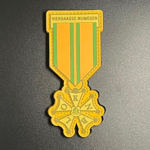 Op de afbeelding: Een gele en oranje medaille met de tekst "VIERDAAGSE NIJMEGEN". De medaille heeft groene strepen en een bloemenontwerp aan de onderkant. Op een zwarte achtergrond.