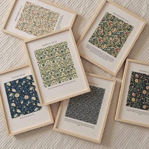May include: A collection of framed art prints featuring floral patterns in various colors, including blue, cream, and green. Each print has a wooden frame and includes a Bible verse. The prints are arranged on a white surface.
