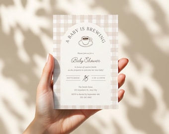 May include: A baby shower invitation held in a hand. The invitation features a beige and white checkered pattern with the text "A Baby is Brewing" and details for the event, including the date, time, and location.