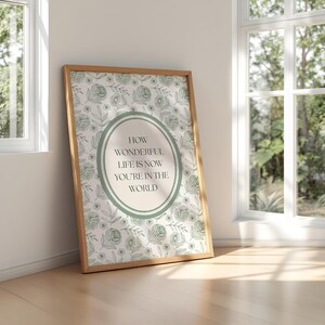 Może przedstawiać: Oprawiony wydruk z kwiatowym wzorem i napisem "How Wonderful Life Is Now You're In The World". Wydruk ma jasnozieloną i kremową kolorystykę, z drewnianą ramą. Wydruk opiera się o ścianę w pobliżu okna.