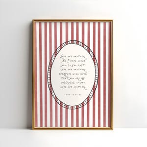 Puede incluir: Una impresión enmarcada con un borde dorado. El fondo presenta rayas verticales rojas y blancas. Un óvalo en el centro contiene texto manuscrito: "Amaos los unos a los otros..." El texto es de Juan 13:34-35.