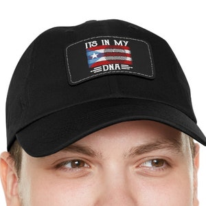 Puede incluir: Una gorra de béisbol negra con un parche blanco que dice "It's in my DNA" y presenta una bandera puertorriqueña.