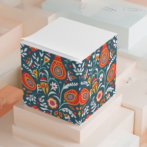May include: A square notepad with a white top and a patterned base. The base has a dark teal background with a floral design in red, orange, yellow, and white. The notepad is on geometric platforms.