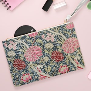 May include: A floral patterned zippered pouch with a white zipper pull and a white wrist strap. The pouch is on a pink surface with a pink notebook, a white pair of sunglasses, a pink lipstick, and a small succulent plant in a white pot.