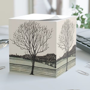 Puede incluir: Un bloc de notas en forma de cubo con una ilustración en blanco y negro de un árbol desnudo en un paisaje. El bloc de notas tiene una hoja superior blanca y un fondo beige.