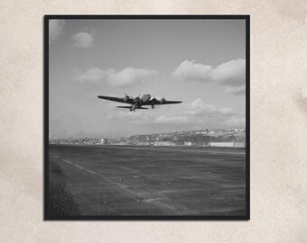 May include: A black and white photograph of a vintage airplane taking off from a runway. The airplane is in mid-air, with its wheels just off the ground. The runway is empty, and there are buildings in the background.