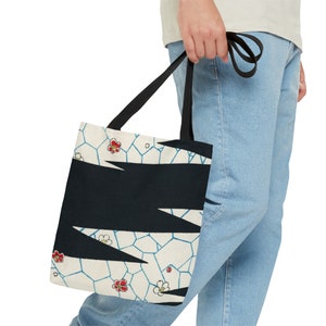 May include: A white tote bag with black geometric shapes and red and white floral designs. The bag has black straps and is being held by a person.