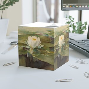 May include: A white water lily with yellow center blooms in a pond with green lily pads. The image is printed on a square notepad.