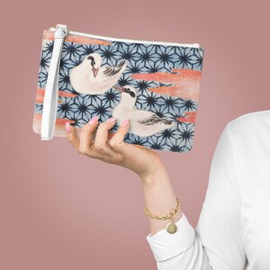May include: A white clutch purse with a blue and white geometric pattern and two white birds with orange accents. The purse has a white strap.