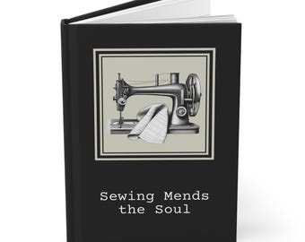 Op de afbeelding: Zwart notitieboek met een ingelijste illustratie van een vintage naaimachine en een gevouwen doek. De tekst "Sewing Mends the Soul" staat in het wit onderaan het notitieboek.