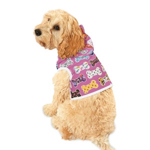 May include: A pink dog raincoat with a white trim and a hood. The raincoat has a bone pattern with different colors and designs. The raincoat is on a light brown dog.
