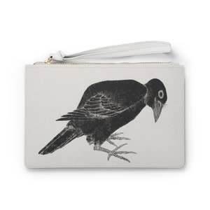 Puede incluir: Una cartera de mano blanca con cremallera con una ilustración en blanco y negro de un cuervo sobre un fondo blanco. La bolsa tiene una correa blanca.