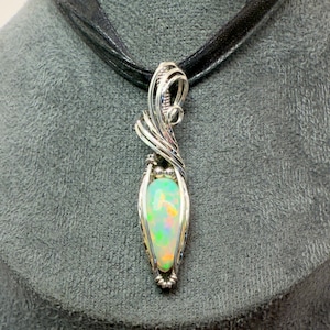 Puede incluir: Un colgante de alambre plateado envuelto alrededor de un ópalo en forma de lágrima con colores iridiscentes. El colgante cuelga de una cinta negra.