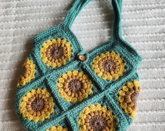 Puede incluir: Una bolsa de mano de crochet con un diseño de girasol. La bolsa está hecha de hilo turquesa, marrón y amarillo. La bolsa tiene dos asas y un botón de madera.