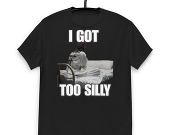 Puede incluir: Camiseta negra con texto blanco que dice "I got too silly". La imagen muestra a una persona con una camisa de fuerza blanca sentada en una cama.
