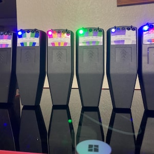 May include: Six black devices with colorful displays and illuminated lights. Each device has a rectangular shape with an arrow design. The devices are arranged in a row on a reflective surface. The text "MARCAUSSINO" is visible on the displays.