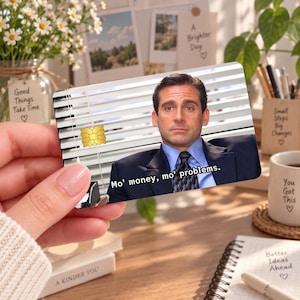 Op de afbeelding: Een rechthoekige creditcardsticker met een afbeelding van een man in een pak en das voor jaloezieën. De tekst Mo' money, mo' problems. staat onderaan in het wit gedrukt. Een goudkleurige EMV-chip is aan de linkerkant zichtbaar.