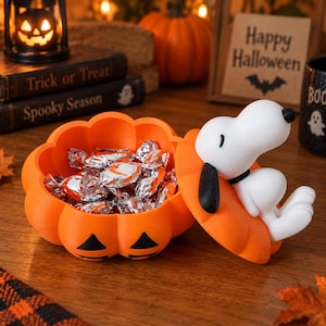 Może przedstawiać: Pomarańczowa miseczka na słodycze w kształcie dyni z pokrywką, na której znajduje się biała figurka Snoopy'ego z czarnymi uszami. Miseczka jest wypełniona cukierkami w srebrnych opakowaniach i ma czarny wzór twarzy dyni z przodu.
