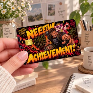 May include: A rectangular credit card skin featuring a vibrant graphic illustration of a man shouting and a cat wearing a crown and sunglasses. The text reads NEEEW ACHIEVEMENT, DUNGEON CRAWLER CARL, and SMALL CAT MASSIVE CHAOS in bold yellow and black.