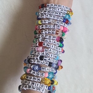 May include: A stack of colorful beaded bracelets with white letter beads spelling out words like "Warrior", "Summoner", "Dragon", "Gun", "Dancer", "Bard", "Reaper", "Scholar", "Knight", "Mage", "Paladin", "Jack", "Ninja", "Sage", "Monk", "Chinist", and "Samurai".