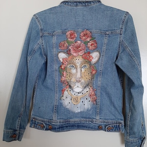 May include: A blue denim jacket with a hand-painted design of a cheetah wearing a crown of pink roses.