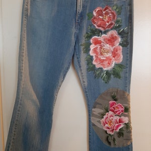 May include: Blue denim jeans with a floral design painted on the right leg. The flowers are pink and white with green leaves. There is a circular patch on the right knee with a camouflage pattern and pink and white embroidered flowers.