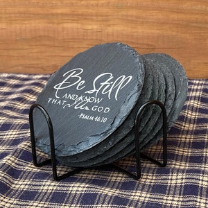 May include: Set of six black slate coasters with a white inscription that reads "Be Still and Know that I am God - Psalm 46:10". The coasters are in a black metal stand.
