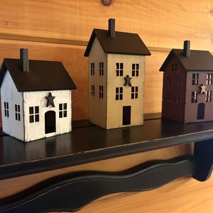 May include: Three small house-shaped decorative ornaments with dark brown roofs and chimneys. The left house is white, the centre is tan, and the right is dark red. Each features a dark star accent on the front facade and multiple cut-out windows.