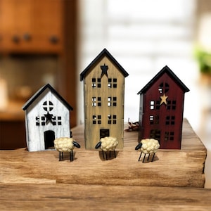 May include: Three decorative house-shaped structures in white, tan, and dark red, each featuring a black roof, cut-out windows, and a star accent. Three small, textured cream-colored sheep figurines with black legs stand in front of the houses.