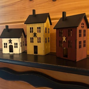 Puede incluir: Tres figuras decorativas de casas de madera con techos y chimeneas negras. La más pequeña es blanca con una estrella negra, la mediana es amarilla con una estrella negra y la más grande es de color rojo oscuro con una estrella color crema.