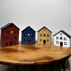 Puede incluir: Cuatro estructuras decorativas de madera en forma de casa con techos negros y una estrella recortada en el centro. Las casas están pintadas en rojo, azul, amarillo y blanco con un acabado desgastado, cada una con ventanas cuadradas y una puerta.