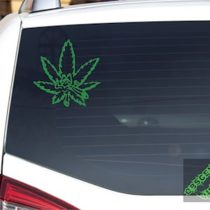 May include: Green vinyl decal of a cannabis leaf with a cartoon face smoking a joint, applied to a car window. The decal is a detailed outline, and the text "OBSCENE VINYL" is in the lower right corner.