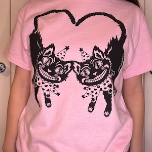 May include: A light pink t-shirt featuring a black graphic of two smiling cat faces with party hats, forming a heart shape. The cats have polka-dotted legs and are set against a plain background.
