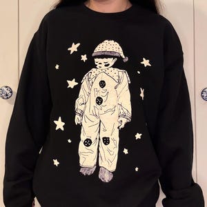 May include: Black sweatshirt featuring a cream-colored illustration of a sad clown with a hat and buttons, surrounded by white stars. The design is a graphic print. The sweatshirt is a casual style.