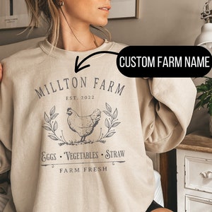 May include: A beige sweatshirt with a graphic design featuring a chicken, leaves, and the text "Millton Farm Est. 2022 Eggs Vegetables Straw Farm Fresh". The text "CUSTOM FARM NAME" is written above the design.