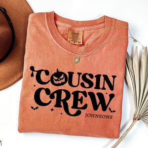 May include: A coral-colored crew neck sweatshirt with the words "Cousin Crew" in black, featuring a jack-o'-lantern, bats, and stars. The name "Johnsons" is printed below. A gold necklace and a brown hat are also visible.