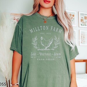 Puede incluir: Una camiseta verde con un dise&ntilde;o gr&aacute;fico blanco que dice "Millton Farm Est. 2022 Eggs - Vegetables - Straw Farm Fresh". El gr&aacute;fico tambi&eacute;n incluye un dibujo blanco de una gallina.