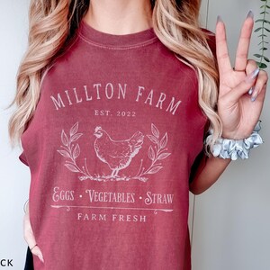 Puede incluir: Una camiseta color granate con un dise&ntilde;o gr&aacute;fico blanco que dice "Millton Farm Est. 2022" con una ilustraci&oacute;n de pollo y el texto "Eggs - Vegetables - Straw Farm Fresh".
