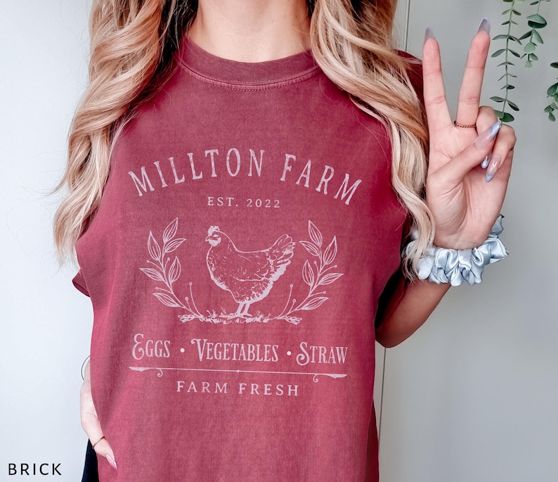 Puede incluir: Una camiseta color granate con un dise&ntilde;o gr&aacute;fico blanco que dice "Millton Farm Est. 2022" con una ilustraci&oacute;n de pollo y el texto "Eggs - Vegetables - Straw Farm Fresh".