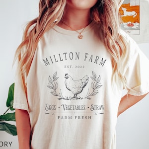 Puede incluir: Una camiseta de color crema con un dise&ntilde;o gr&aacute;fico en blanco y negro. El gr&aacute;fico presenta una gallina, hojas y el texto "Millton Farm Est. 2022 Eggs Vegetables Straw Farm Fresh".