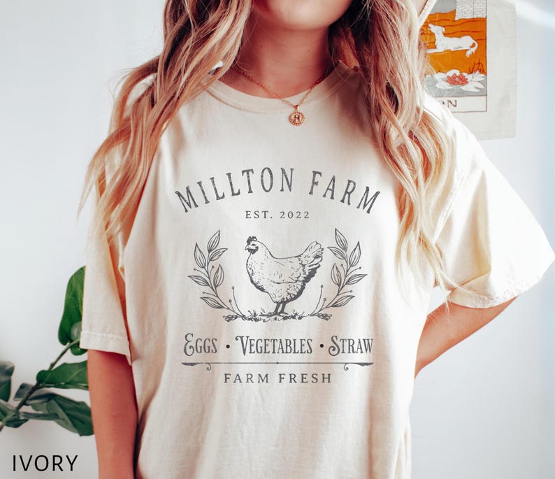 Puede incluir: Una camiseta de color crema con un dise&ntilde;o gr&aacute;fico en blanco y negro. El gr&aacute;fico presenta una gallina, hojas y el texto "Millton Farm Est. 2022 Eggs Vegetables Straw Farm Fresh".